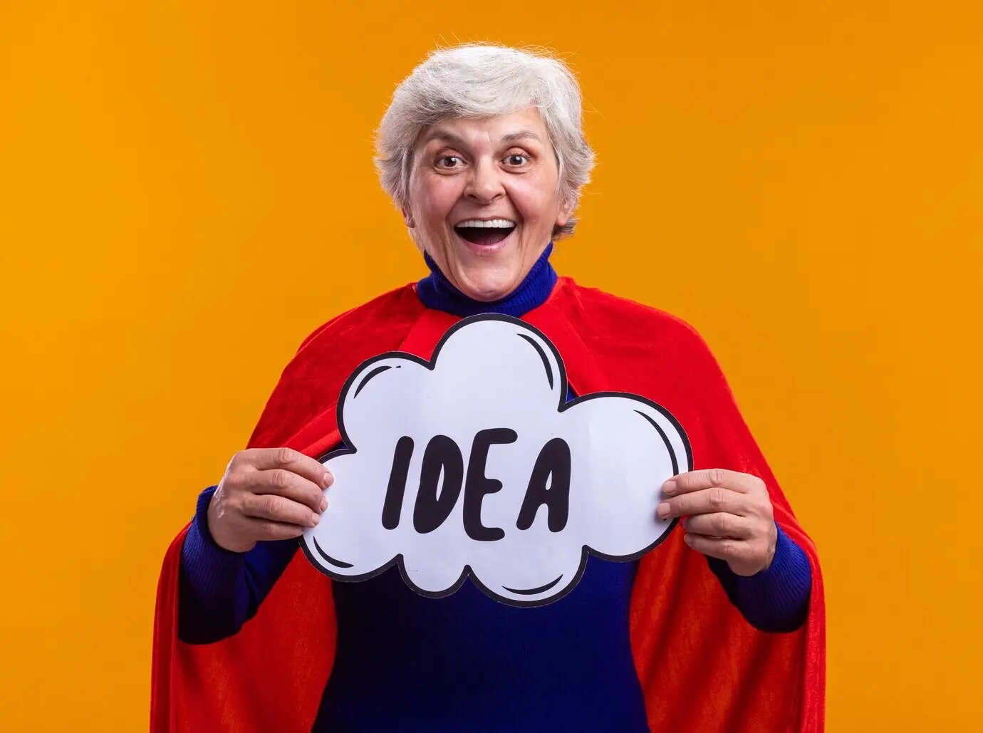 Eine glückliche, begeisterte Seniorin als Superheldin mit rotem Umhang hält ein Sprechblasenschild mit dem Wort „idea“, blickt in die Kamera und lächelt fröhlich; sie steht vor einem orangefarbenen Hintergrund.